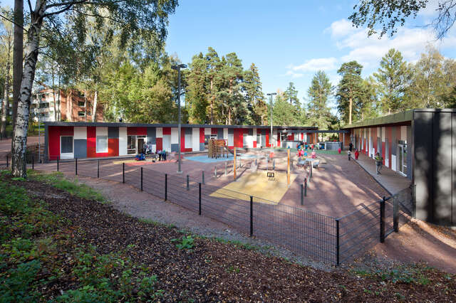 Day care centre consisting of two wings with red, white and grey panels covering the facade with a playground in front of the building.