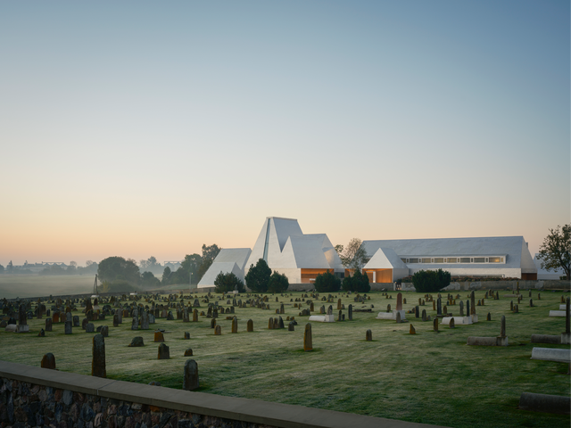 Hazy photograph of a cemetery, with an aluminium building in the background featuring pitched roofs of varying heights.