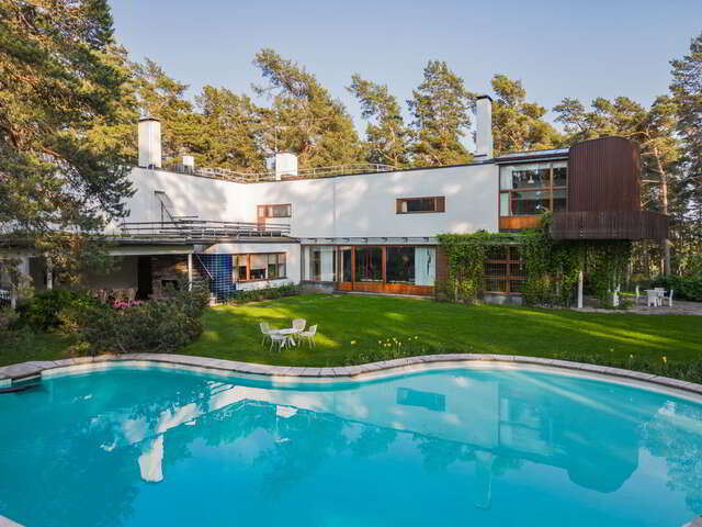 A freeform pool in a green garden, white rendered house with wooden parts in the background