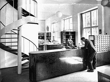 A librarian and visitor at a desk, a spiral staircase to the left.