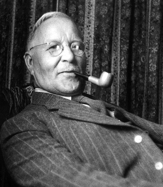 An elderly man in a suit leaning back in a chair with a pipe in his mouth