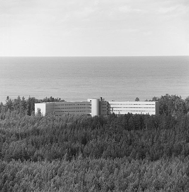 Birds eye view of the white hotel building surrounded by trees and with the sea in the background.