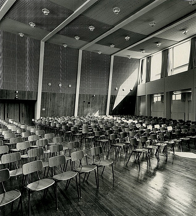 Black and white picture of the school hall filled with rows of wooden chairs.