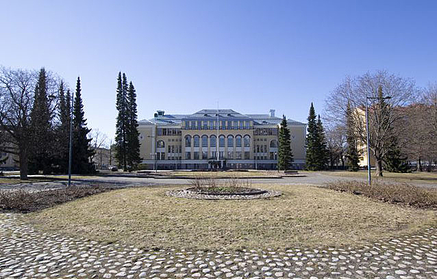Today's facade of the building with white, yellow and brown elements viewed from the other side of the park.