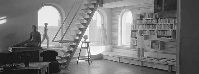 Black and white image of a light wooden interior with arch windows, female sculptures and a bookshelf with a wooden ladder leading upstairs.