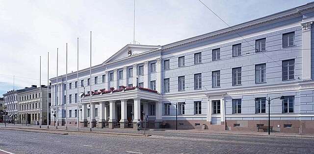 Long light blue building with a white entrance supported by columns that also works as a balcony for the second floor.