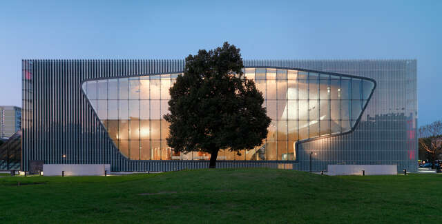 Box-shaped building with a giant window, big tree on a green hill in the foreground