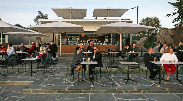 People sitting at an outdoor café under white umbrellas. Paved, dark floor pavement in the foreground, open bar counter in the background.