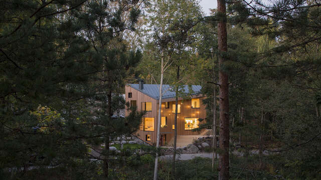 A wooden detachde house in the forest, with a non-treated wood facade and light shining from the windows