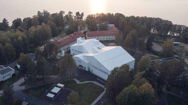 An aerial view of a large white building mostly surrounded by trees. A Large body of water can also be seen.