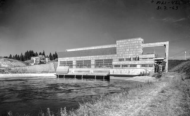 Black and white photo of the power plant, displaying the power plant in function. The water of the river pours out of the power plant back into the river.