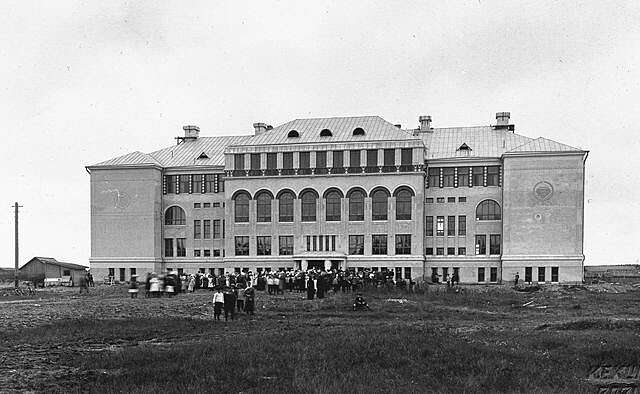 Black and white picture of the old school facade with a large group of people in front of it.