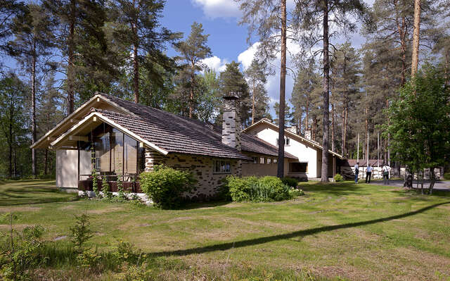 A house with a hipped roof laid from red bricks. The house has a chimney that is decorated with a beautifully, organically laid white and dark tiles. In the front there is a glazed porch facing the sun.