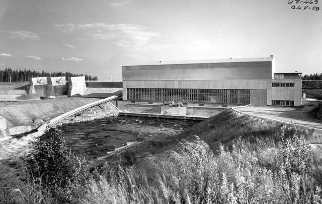 Image of hydropower plant seen from the river. The facade is covered with metal sheets and brick. Ribbon windows let the light in.