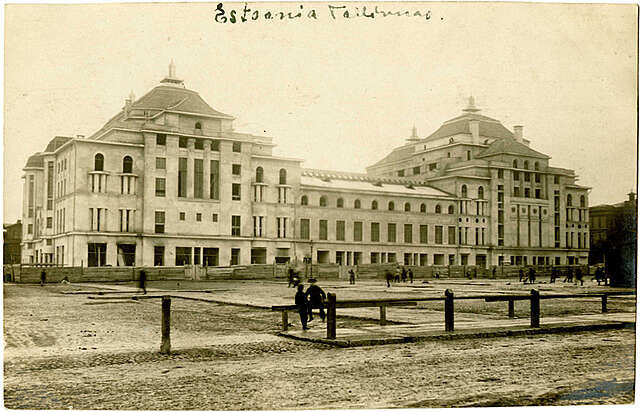 Old black and white picture of a grand building with people walking in the square in front of it.