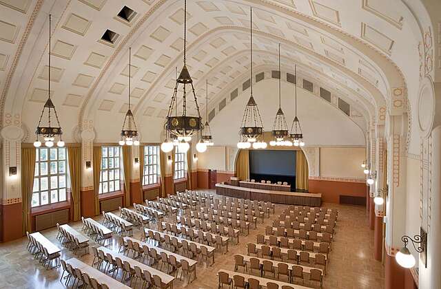 Large assembly hall with white ornate walls with peach colored details, large grid windows framed by mustard yellow curtains and metal light fixtures hanging from the arched ceiling.