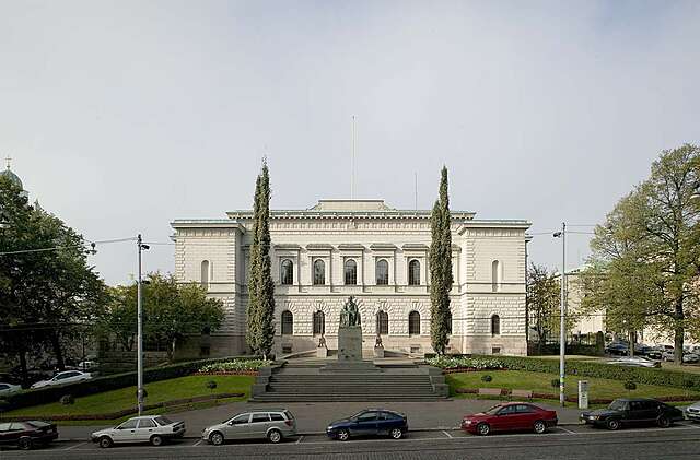White facade of the bank of Finland with trees in both sides of the entrance and cars lined up on the street.
