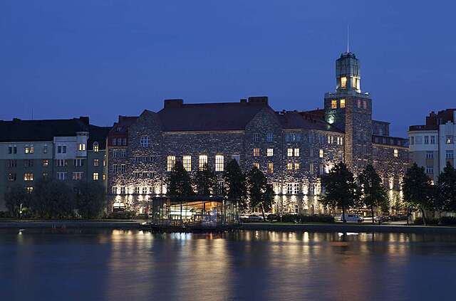 Old stone building photographed at night with the lights on, in the from there is a small glass building on the shore.
