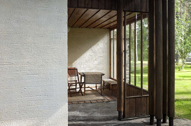 Photo showcasing the veranda of the house and how the sunlight can access the veranda beautifully. The walls of the veranda are decorated with warm wooden columns. The flooring has a wooden blank surface as well as a stone flooring next to it. The materials feel rich and diverse, and they possess this initial feeling of warmth.