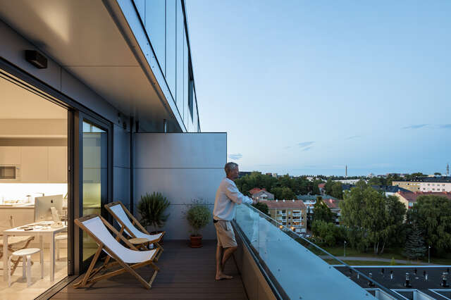 A person standing on a balcony of an apartment, looking out at a city skyline during sunset.