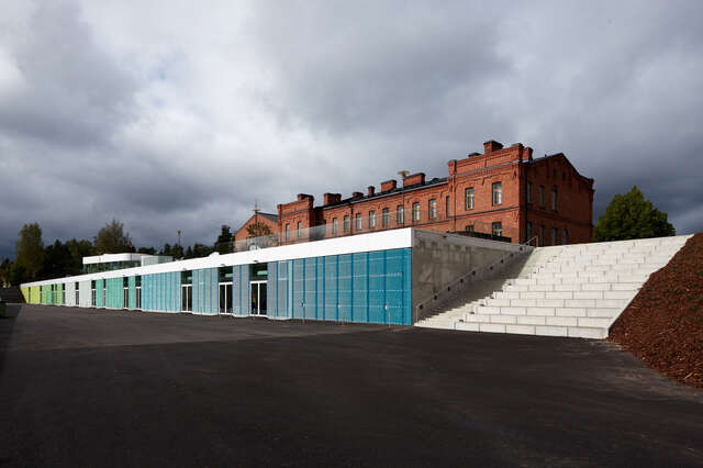 Square annex with blue-green-turquoise steel grid façade and white stairs, red brick barracks in the background