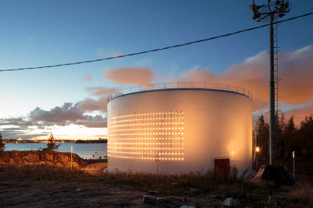 White oil silo surrounded by nature and water. Its walls are perforated with 2012 holes.