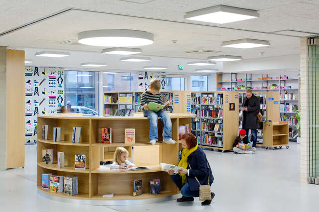 Children and adults in a luminous school library reading books in the middle of the book shelves