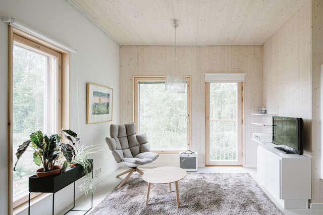 Interior picture of one flat with wooden walls, two large windows and one french window. Decorated with light materials, forested landscape showing from the windows.
