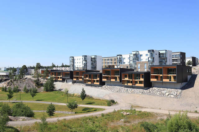Row of wooden houses with large apartment building complex behind it. Grass field in front of wooden houses