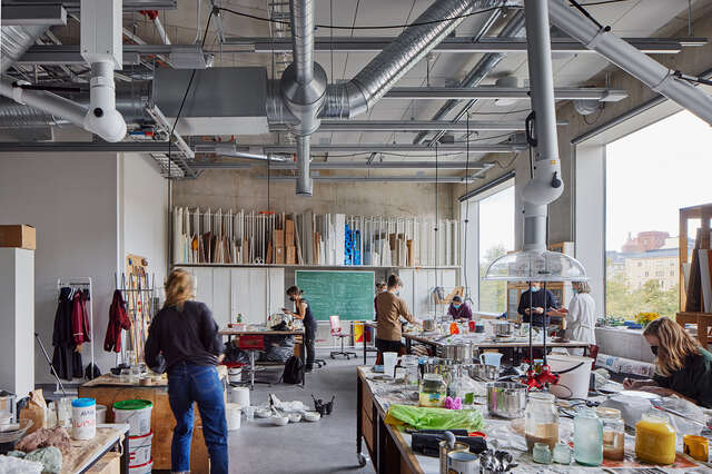 Room with large tables full of art supplies. 8 people in the room with facemasks on. Two large square windows on right side wall.