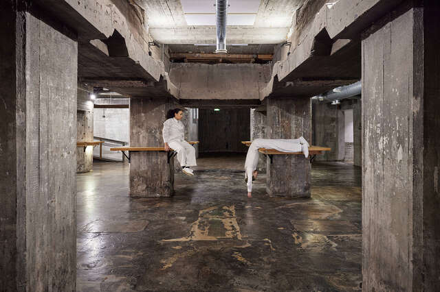 Old and worn looking room made of concrete with wooden tables attached to pillars. Two people in white clothing. sitting on table tops.