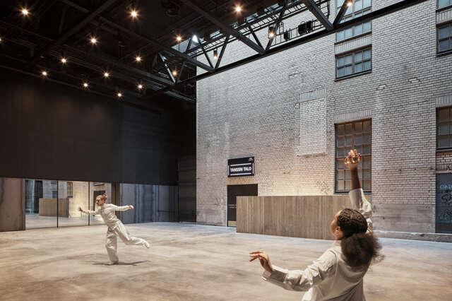 Large indoor space with two people dancing. Large white brick wall to the right. Multiple small lights on ceiling.