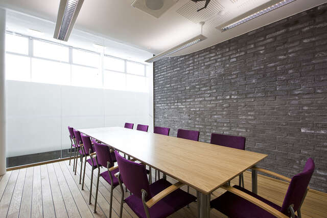 Meeting room with a long table and purple chairs, there is a glass- and a dark brick wall.