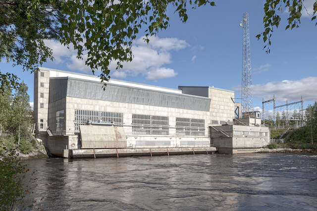 Photo that shows the power plant over the river during the day. The façade is divided into three masses, one main horizontal mass in the front with three main rectangular windows.