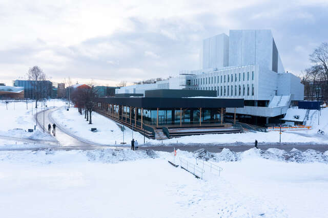 Wooden structure surrounded by snow. Large white building to the right.
