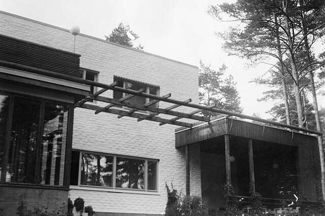 White exterior with a protruding living room and a wooden grid hanging between it and the entrance.