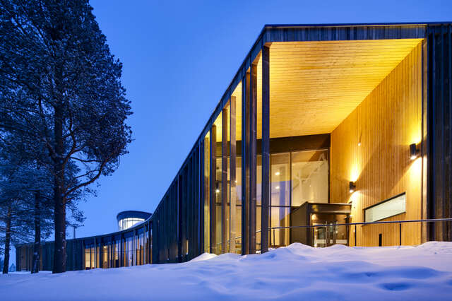 Entrance with high ceiling, wooden details and orange light, snow on the ground and evening sky.