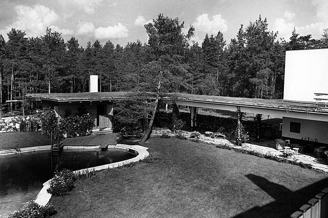 Black and white picture of the garden with a pool, the elongated roof covering the terrace in in the back.