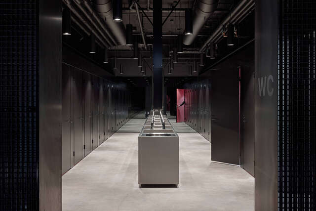 Marge bathroom with black stall doors on both sides. Large metal washing area in the middle. Concrete floor.