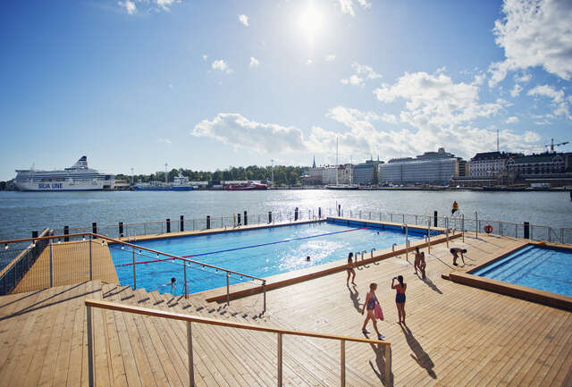 Swimming pool surrounded by a wooden terrace, next to the ocean. In the background there e is the city shoreline and. a cruise ship.