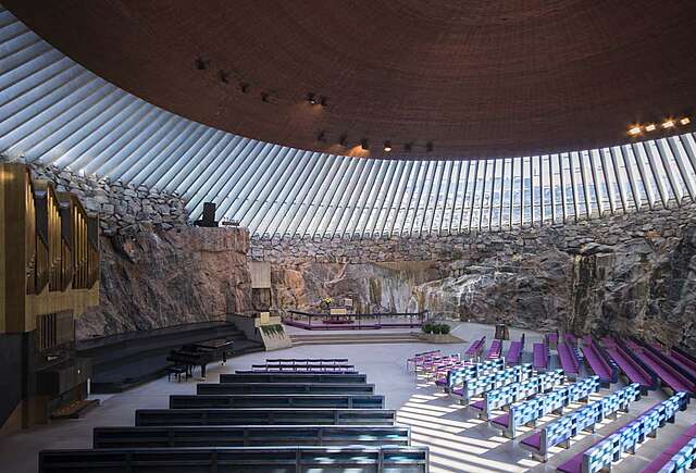 Interior of the rock church with purple cushioned bench rows, a window panel surrounding every side of the room and a wooden rounded ceiling on top