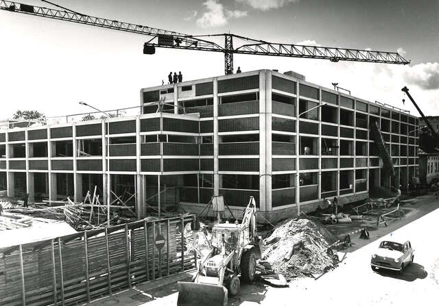 Black and white picture of the construction site with an almost ready building, a crane in the background and a digger in the foreground next to a large pile of sand.