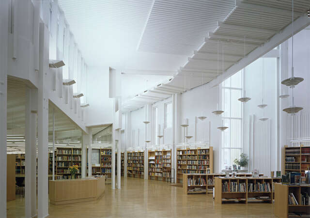 Spacious library interior with a information desk a t the left, wooden bookshelves and pendant lights.