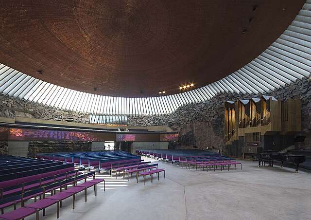 Interior of the rock church with purple cushioned bench rows, a window panel surrounding every side of the room and a wooden rounded ceiling on top.