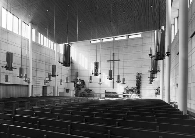 Black and white photo of a high space with a band of windows below the ceiling. Rows of benches, cross on the wall