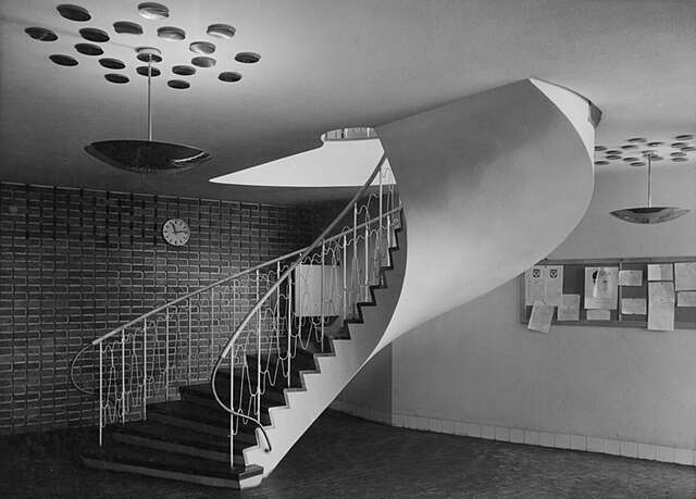Spiral stairs with white metal railings in a room with a notice board on the right side and a brick wall on the left.