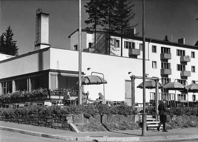 Commercial premises in front of a block of flats, a man passing by, women sitting on the terrace