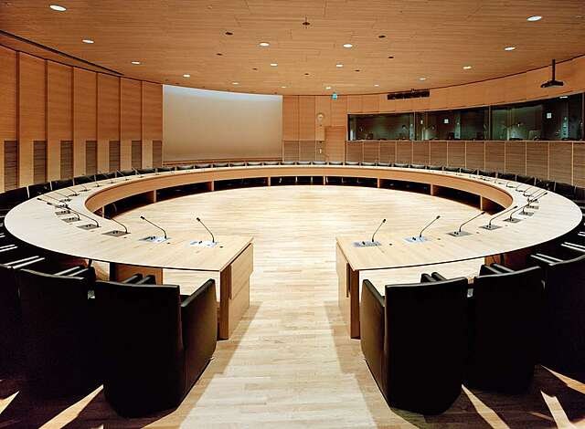Black armchairs arranged in a circle with a circular table with microphones on every seat. In the middle of the room there is a circular open area.