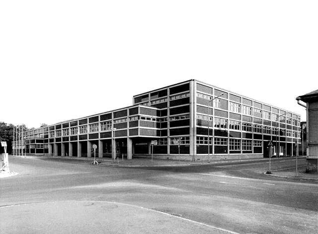 Black and white picture of the building facade. The right side has three floors while the left side has only two along with a protruding extension on the second level which is supported by pillars.