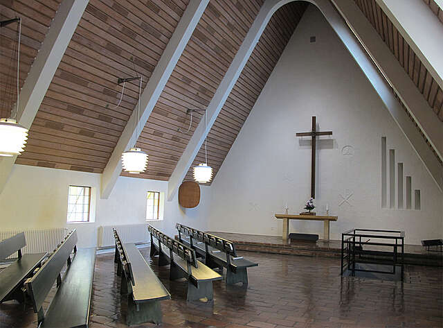 Chapel with white walls, a gable roof with wooden paneling and white support beams on the sides, the square windows with stained glass give the space a comfortable feel.